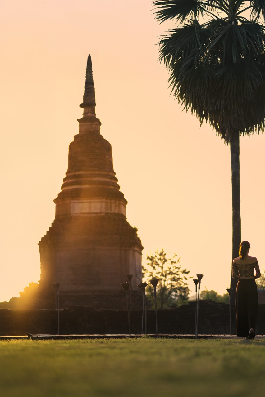 Ancient Sukhothai temple stupa silhouetted at sunset with torches and a palm tree